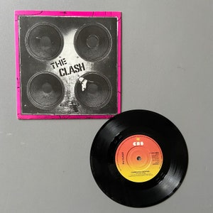 Könnte beinhalten: Eine schwarze Schallplatte mit einem roten, orangen und gelben Etikett. Das Etikett trägt den Text "CBS" in einem weißen Kreis, "The Clash" in schwarzem Text und "Complete Control" in schwarzem Text. Die Schallplatte liegt auf einer grauen Oberfläche.