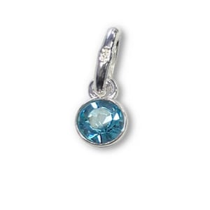 May include: A small, round, light blue gemstone pendant with a silver-colored bail. The gemstone is set in a silver bezel and has a faceted surface. The pendant is designed to be worn as a necklace charm.