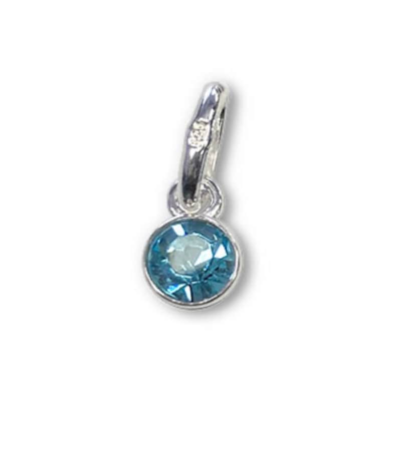 May include: A small, round, light blue gemstone pendant with a silver-colored bail. The gemstone is set in a silver bezel and has a faceted surface. The pendant is designed to be worn as a necklace charm.