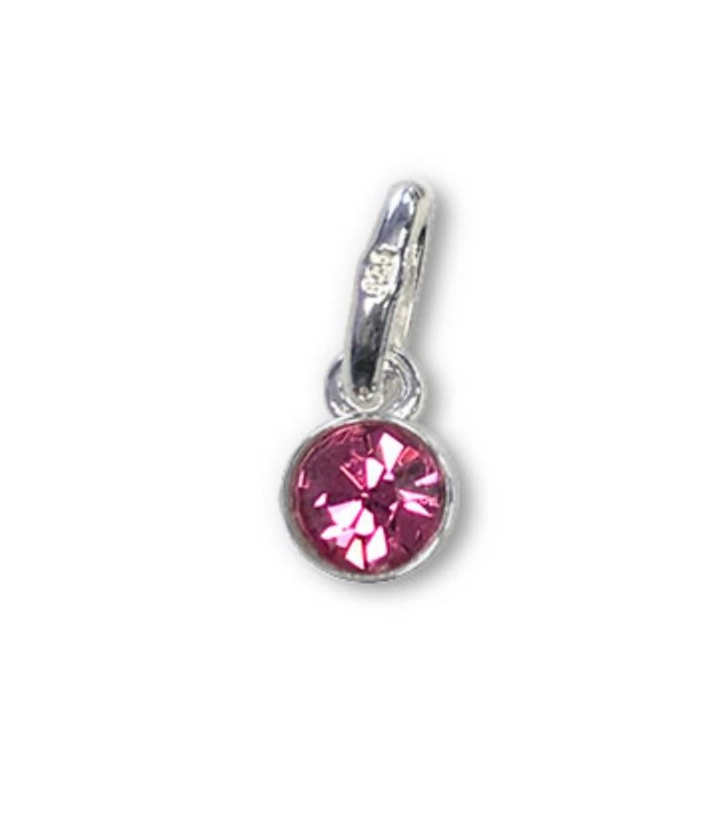 May include: A small, round, pink gemstone charm with a faceted surface, set in a silver-colored metal bezel. The charm has a loop at the top for attaching to a necklace or bracelet. The background is plain white.