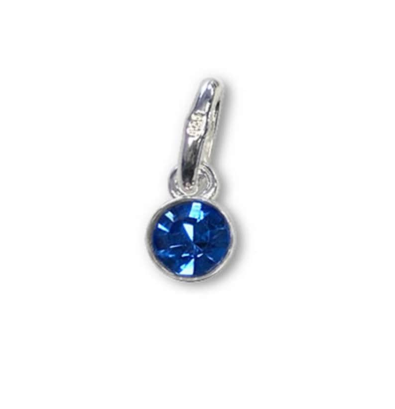 May include: A small, round, blue gemstone charm with a silver-colored bail. The charm has a faceted, sapphire-like appearance, and is suspended from a simple, polished metal loop. The background is plain white.