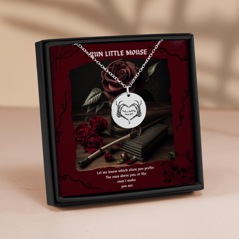 Peut inclure: Un collier en argent avec un pendentif rond repr&eacute;sentant un c&oelig;ur form&eacute; de mains squelettiques. Le pendentif porte l'inscription "Pick a fighting mood girl?" Le collier est pr&eacute;sent&eacute; dans une bo&icirc;te noire avec un fond rouge et un motif de rose. La bo&icirc;te porte &eacute;galement l'inscription "Run Little Mouse".