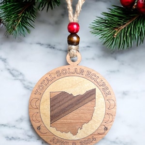 May include: A wooden ornament shaped like the state of Ohio with the text "TOTAL SOLAR ECLIPSE APRIL 8, 2024" around the outside. The ornament is brown and has a natural wood grain.