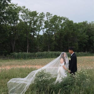 May include: A wedding dress with an off-the-shoulder neckline and a long, flowing white veil, paired with a black suit and white shirt. The attire is worn by two individuals standing together in a grassy field with a line of tall green trees behind them.