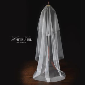 May include: A white tulle wedding veil with two layers. The veil is draped over a mannequin and is displayed against a black background.