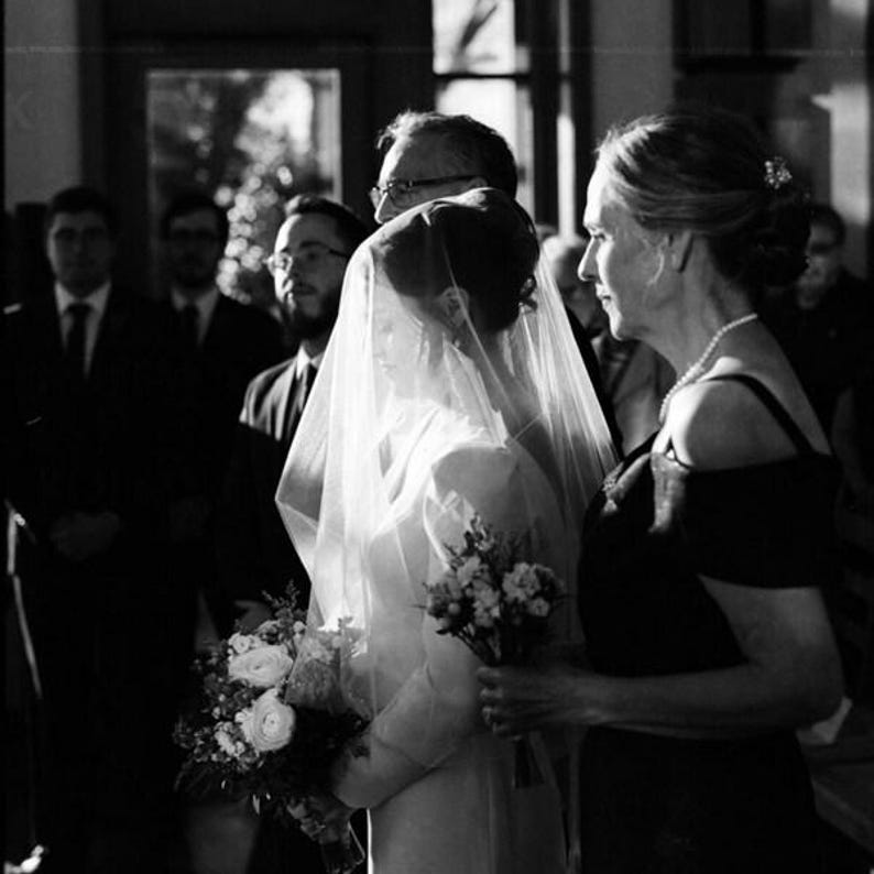 May include: A black and white photograph of a person wearing a wedding veil over their face, holding a bouquet of flowers. Beside them, another person wears a dark dress and a pearl necklace, also holding a bouquet. Guests stand in the background.