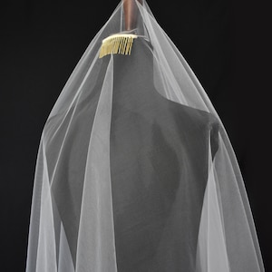May include: A sheer white tulle veil attached to a gold-toned metal hair comb. The lightweight fabric drapes downward in soft, vertical folds, creating a layered appearance. The comb is positioned at the top, securing the translucent material.