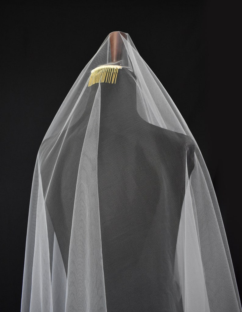May include: A sheer white tulle veil attached to a gold-toned metal hair comb. The lightweight fabric drapes downward in soft, vertical folds, creating a layered appearance. The comb is positioned at the top, securing the translucent material.