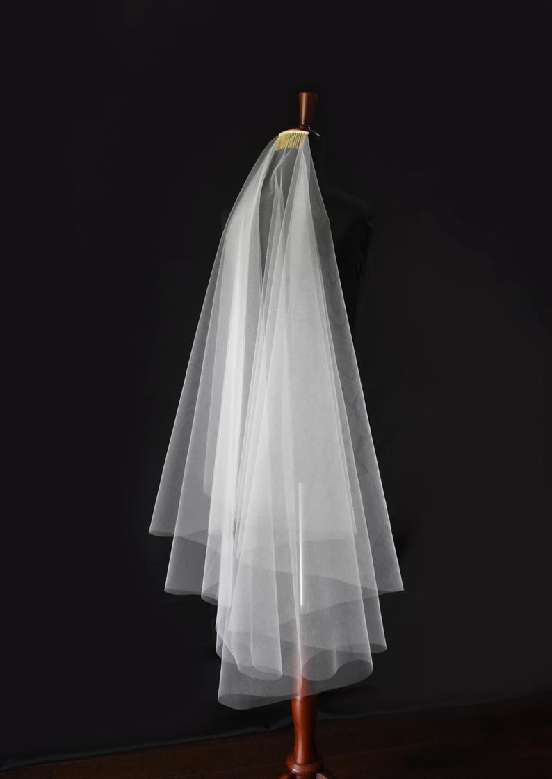 May include: A sheer white wedding veil made of fine tulle fabric, featuring multiple cascading layers that create a soft, voluminous silhouette. The veil is securely attached to a gold-toned metal comb, designed for easy placement in hair.