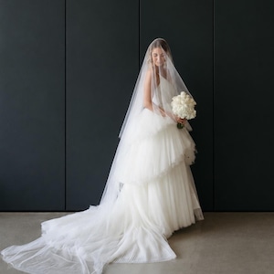 May include: A white floor-length wedding dress with a tiered tulle skirt and a long, sheer veil draped over the head. The dress features a sleeveless bodice and a full, flowing train that extends across the floor. A bouquet of white roses is held.