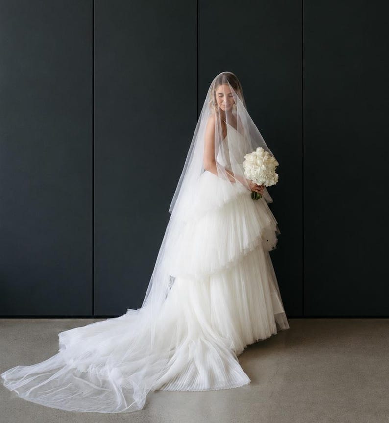 May include: A white floor-length wedding dress with a tiered tulle skirt and a long, sheer veil draped over the head. The dress features a sleeveless bodice and a full, flowing train that extends across the floor. A bouquet of white roses is held.