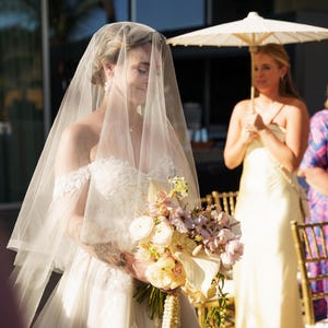May include: An off-the-shoulder white wedding dress with lace detailing, a sheer veil covering the head, and a bouquet of white, cream, and light pink flowers. A person stands in the background holding a white parasol over their shoulder.