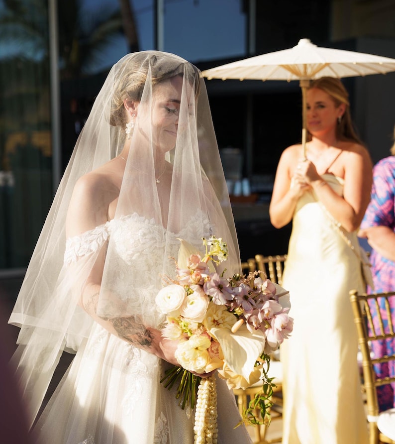 May include: An off-the-shoulder white wedding dress with lace detailing, a sheer veil covering the head, and a bouquet of white, cream, and light pink flowers. A person stands in the background holding a white parasol over their shoulder.