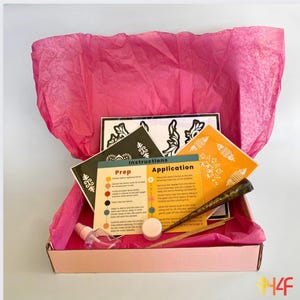 May include: A pink box filled with supplies for creating henna tattoos. The box contains two stencils, a cone of henna paste, a small bottle of solution, a small jar of henna paste, and instructions for prepping and applying the henna.