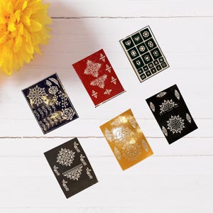 May include: An overhead shot displays six rectangular henna stencils in various colors, including red, black, and gold. Each stencil features intricate white floral and geometric designs. A yellow flower is in the upper left corner.