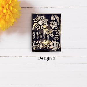 May include: A black stencil sheet with gold-colored designs of flowers, leaves, and a snowflake. The text "Design 1" is printed below the stencil.