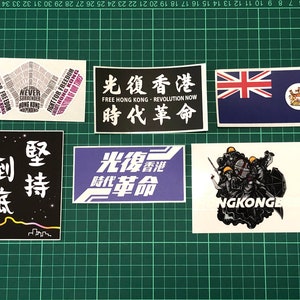 Puede incluir: Cinco pegatinas con diferentes diseños y textos. La primera pegatina es una nube de palabras rosa y morada con el texto "Fight for Freedom" y "Liberate Hong Kong". La segunda pegatina es negra con texto blanco que dice "Free Hong Kong - Revolution Now" y "時代革命". La tercera pegatina es una bandera azul y blanca con una cruz roja y una corona. La cuarta pegatina es negra con texto blanco que dice "光復香港" y "堅持到底". La quinta pegatina es blanca con una ilustración en blanco y negro de cuatro personas con cascos y armas. El texto "NGKONGE" está debajo de la ilustración.