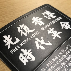 May include: Black and white sticker with the text "Free Hong Kong - Revolution Now" and the Chinese characters "光復香港" and "時代革命".