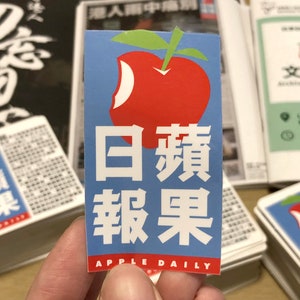 Puede incluir: Una pegatina azul con una manzana roja con un mordisco. El texto "日蘋 報果 APPLE DAILY" está impreso en la pegatina.
