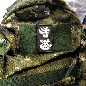 Puede incluir: Una mochila de camuflaje verde con un parche negro que dice "台灣" en caracteres chinos blancos.