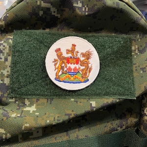 May include: A white circular patch with a gold embroidered coat of arms featuring a lion, a dragon, and a shield with a red maple leaf. The text "HONG KONG" is written in a blue circle around the coat of arms.