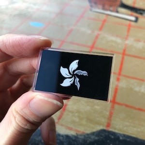 May include: A black and white badge featuring a stylised white flower design. The badge is made of clear plastic and has a black background.