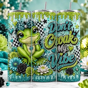 May include: A cylindrical tumbler with a straw, featuring a green frog with a teal leopard print bow, surrounded by teal and black flowers, checkered patterns, and glittery drips. The text reads Don't Croak My Vibe in glittery teal and green.