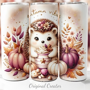 Op de afbeelding: Een witte beker met een herfstthema. Het heeft een schattige egel die een kopje koffie vasthoudt, omringd door pompoenen, bladeren en de tekst "autumn vibes". Het ontwerp is in tinten oranje, paars en bruin.