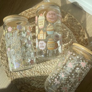 May include: Three glass tumblers with bamboo lids and straws. One tumbler has the text "Sabr" with floral accents. Another has the text "Sabr is Sweet" and other graphics. The third has a floral pattern with bows.