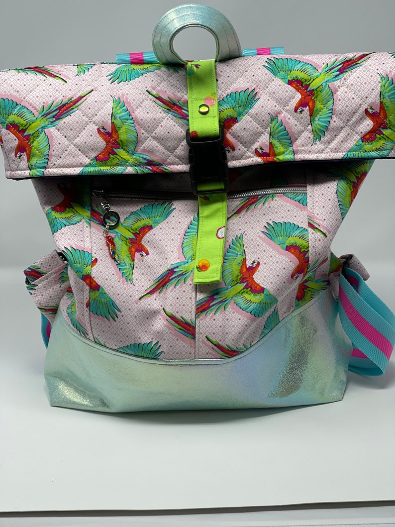 Puede incluir: Una mochila acolchada rosa y verde con estampado de loros. La mochila tiene una solapa con cierre de hebilla y un fondo azul iridiscente.
