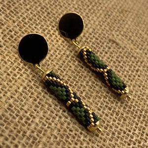 May include: A pair of gold-toned earrings with black circular studs. The earrings feature cylindrical beaded pendants in green, black, and gold patterns. The earrings are displayed on a textured, tan surface.