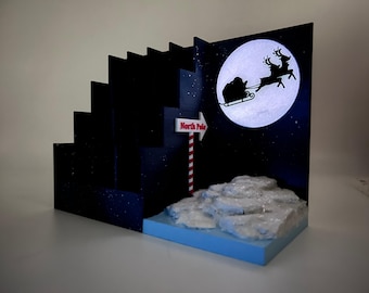 Può includere: Un espositore blu scuro a più livelli con la sagoma di Babbo Natale su una slitta trainata da renne davanti a una grande luna bianca. Un cartello a strisce rosse e bianche indica il Polo Nord, sopra una base di neve bianca testurizzata e glitterata.