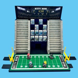 May include: A Bitty Bowl football stadium display stand featuring a scoreboard with the text Bitty Bowl, two sets of grey tiered bleachers, a green field with white yard lines and goalposts, and twelve small collectible figures in boxes.