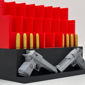 May include: A tiered red storage rack with multiple vertical slots for ammunition, featuring a row of gold-colored bullet replicas. In front, a black base holds two grey, textured pistol models positioned at an angle, displaying detailed grips and slides.