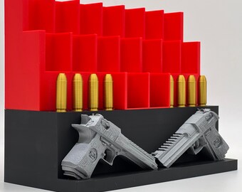 May include: A tiered red storage rack with multiple vertical slots for ammunition, featuring a row of gold-colored bullet replicas. In front, a black base holds two grey, textured pistol models positioned at an angle, displaying detailed grips and slides.