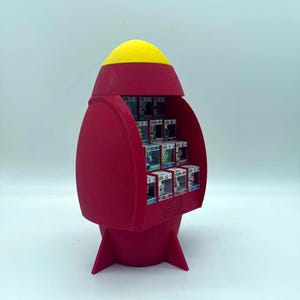 Può includere: Un espositore a forma di razzo rosso con punta gialla, dotato di diversi ripiani interni che contengono piccole scatole quadrate con finestre trasparenti. La base del razzo presenta tre pinne ed è progettata per contenere delle statuette.