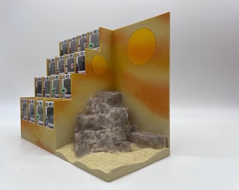 Peut inclure: Un présentoir à gradins pour figurines de collection avec un thème de paysage désertique. La structure présente un design en escalier avec un fond peint en orange et jaune, deux soleils circulaires, une base sablonneuse et des rochers gris.