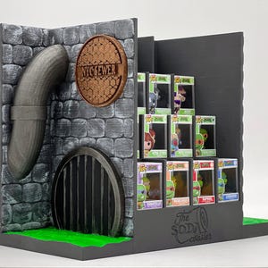 Peut inclure: Un présentoir avec un mur en pierre grise, un tuyau courbé, une plaque d'égout circulaire NYC SEWER et une grille noire. Le support contient plusieurs figurines en boîte et porte l'inscription The Soda Coaster sur la base.