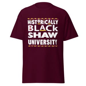 Peut inclure: Un t-shirt bordeaux avec un texte blanc qui dit "Historically Black Shaw University" avec un motif g&eacute;om&eacute;trique jaune et rouge au-dessus et en dessous du texte.