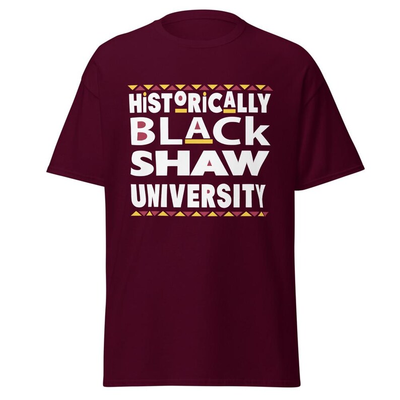 Peut inclure: Un t-shirt bordeaux avec un texte blanc qui dit "Historically Black Shaw University" avec un motif g&eacute;om&eacute;trique jaune et rouge au-dessus et en dessous du texte.