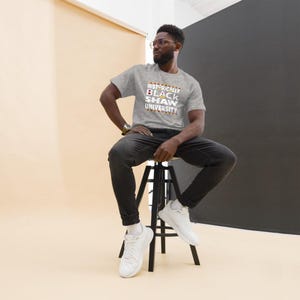 Peut inclure: Un homme portant un t-shirt gris avec le texte "Historically Black Shaw University" en lettres blanches et orange. Il est assis sur un tabouret noir et porte un jean noir et des baskets blanches.