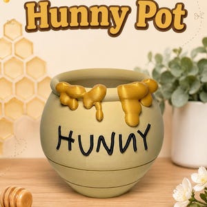 May include: A 3D printed tan pot with a rounded shape, featuring a textured golden yellow honey drip design around the top rim and the word HUNNY written in black letters on the front. The pot has a smooth finish and a wide opening at the top.