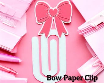 May include: A white paper clip with a large pink bow and a small pink heart at the center. The text Bow Paper Clip 3MF/STL is printed in black at the bottom. The object is surrounded by various pink stationery items on a solid pink background.