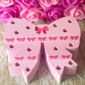 May include: A light pink bow-shaped decorative item with small bow accents and circular cutouts. The background features a wall of pink and magenta roses, with the text "@ Nurse Ronda Creations" in glittery pink.