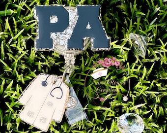 May include: A badge reel with the letters "PA" in navy blue with silver glitter accents. The reel features a white lab coat charm with a stethoscope and the text "Nurse Ronda Creations". The background is green grass with clear diamond-shaped decorations.