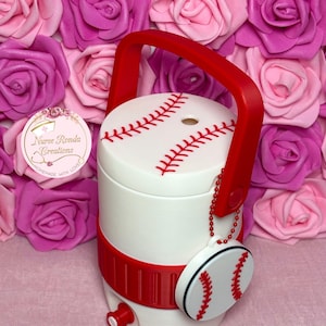 May include: White and red baseball-themed cooler with a red handle and a baseball charm. The cooler has red stitching details and a red band around the middle. The charm is a miniature baseball. The cooler is decorated with a baseball design.