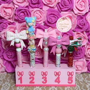 May include: Five decorative pens with various toppers including bows and a character figure, displayed in a pink rectangular stand labeled with numbers one through five and bow icons. The stand features the text Nurse Ronda Creations.