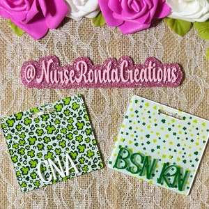 May include: Two green and white patterned plastic badge holders with the text "CNA" and "BSN RN" in white lettering. The badge holders are on a brown burlap background with pink glitter letters that read "@NurseRondaCreations".