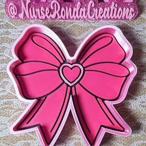 Könnte beinhalten: Ein rosa schleifenförmiger Ausstecher mit einem Herz in der Mitte. Die Schleife hat schwarze Umrisse und einen weißen Rand. Der Text "@NurseRondaCreations" befindet sich in rosa Glitzer über der Schleife.