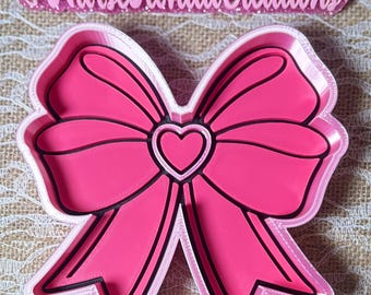 May include: A pink bow-shaped cookie cutter with a heart in the center. The bow has black outlines and a white border. The text "@NurseRondaCreations" is above the bow in pink glitter.